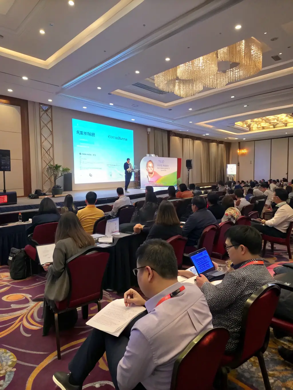 A modern conference hall filled with attendees listening to a keynote speaker on a large screen, focusing on the energy and engagement of the audience.